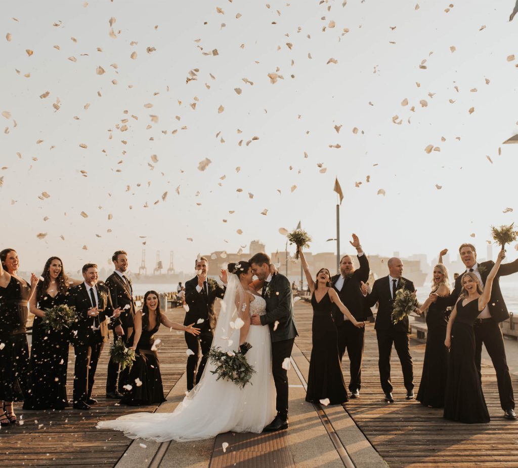 A wedding party cheers and throws confetti around the couple on a waterfront dock