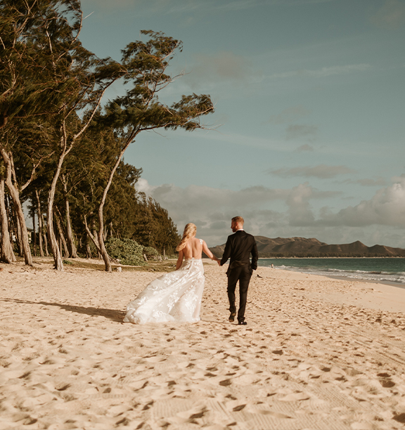 A bride and groom walk along a tree-lined beach in Hawaii