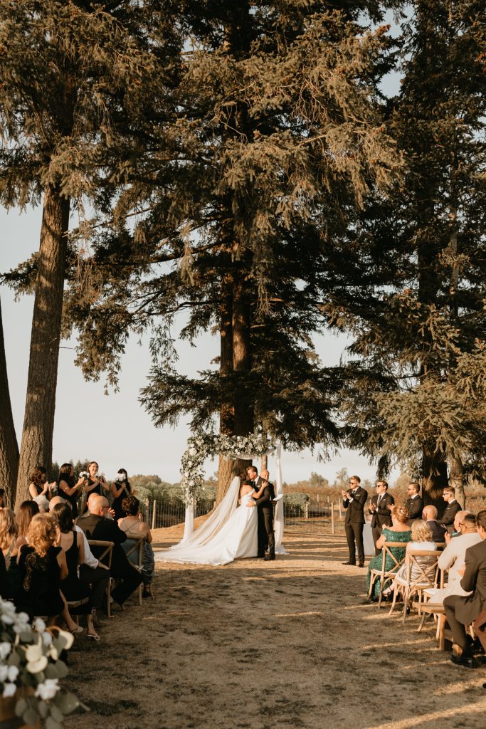Guests line a tree-shaded aisle as a couple says their vows outdoors