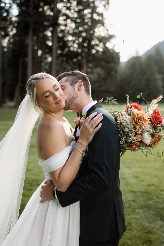 A bride and groom share a kiss, her bouquet of orange flowers in hand, with mountains behind them