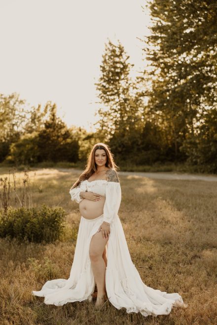 Stunning mama in elegant dress walking through sunlit meadow during maternity shoot
