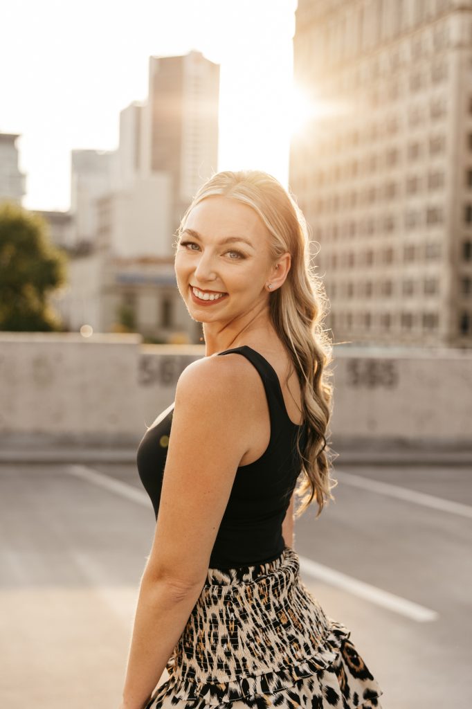 A smiling woman in a black top looks over her shoulder on a city rooftop at sunset