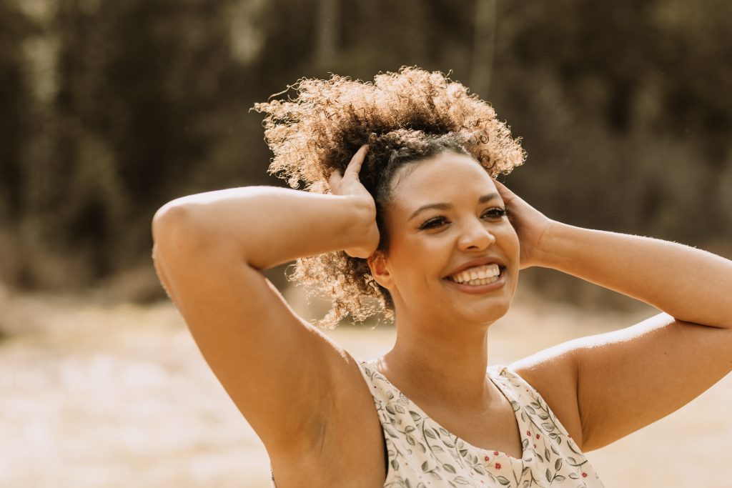 A woman with curly hair laughs, both hands in her hair, in warm golden light
