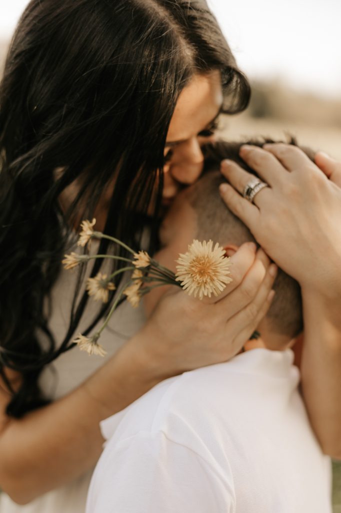 A mother presses a kiss to her child's head, a dried wildflower in her hand