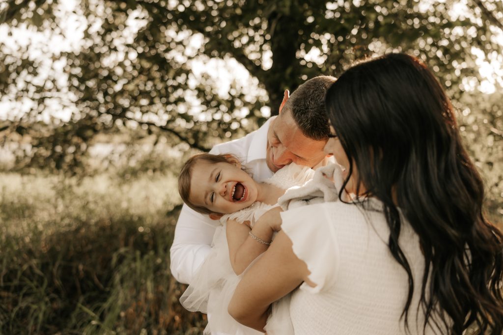 Parents hold their laughing toddler between them under a leafy tree