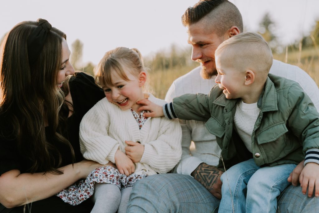 A family of four sits close together in a field, everyone laughing