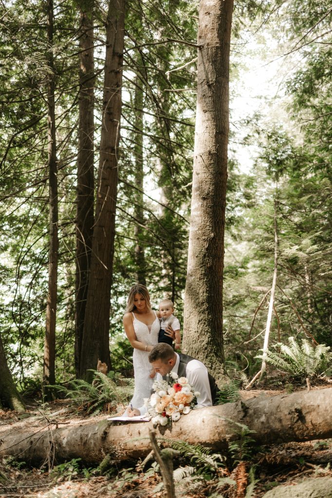 A couple signs their marriage licence on a fallen log in a tall green forest