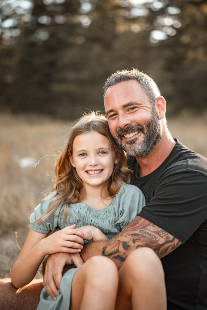 A smiling dad sits in the grass with his daughter on his knee
