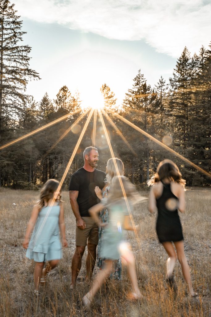 A family walks and twirls through a meadow in Chilliwack as the sun bursts through the trees behind them