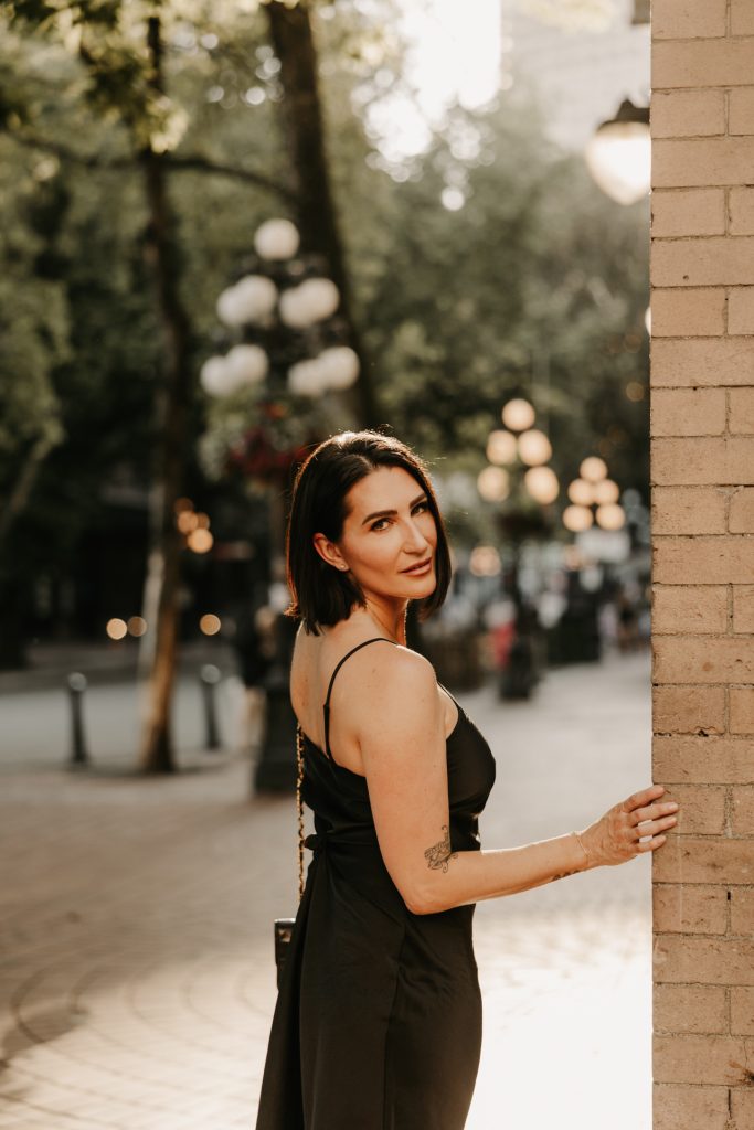 A woman in a black dress leans against a brick wall on a Gastown street lit by old lamps