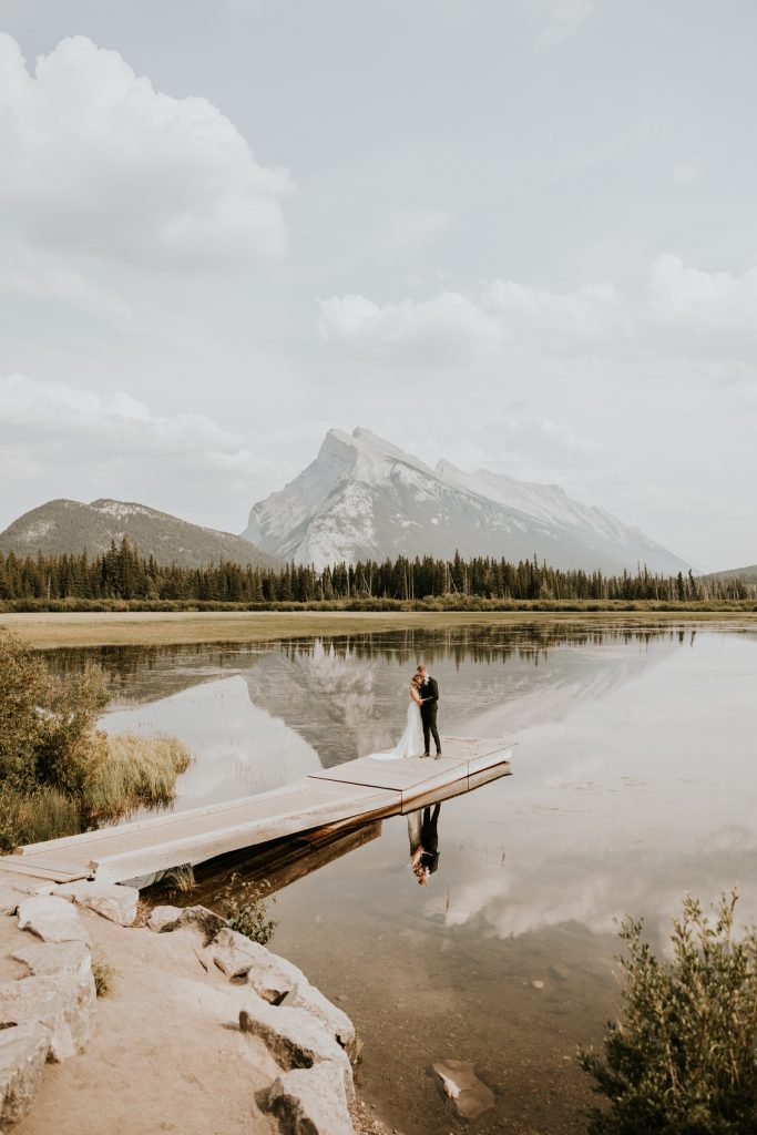 Bride and groom embracing on a wooden dock on a still mountain lake in Banff, Alberta, the peak reflected in the water