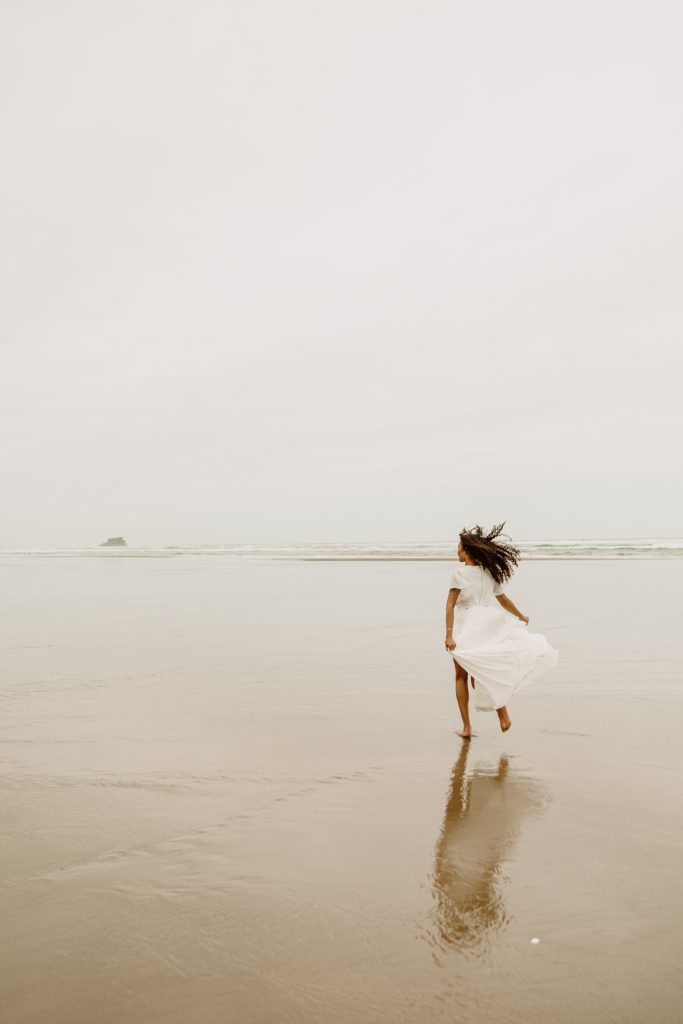 A woman in a white dress runs across wet sand toward the sea