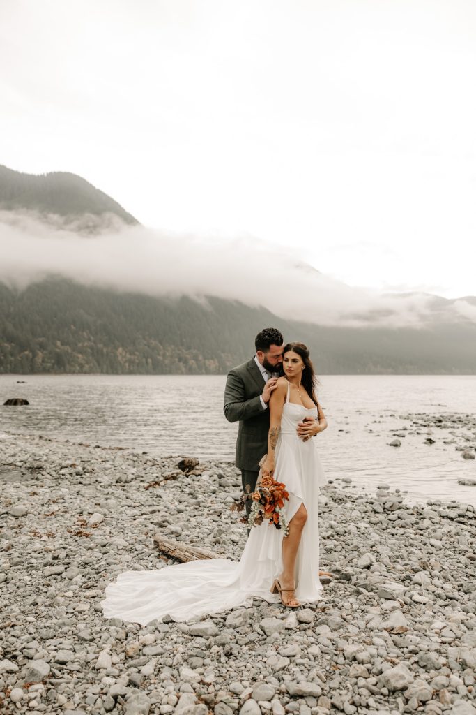 A couple embraces on a pebbled lakeshore beneath misty mountains
