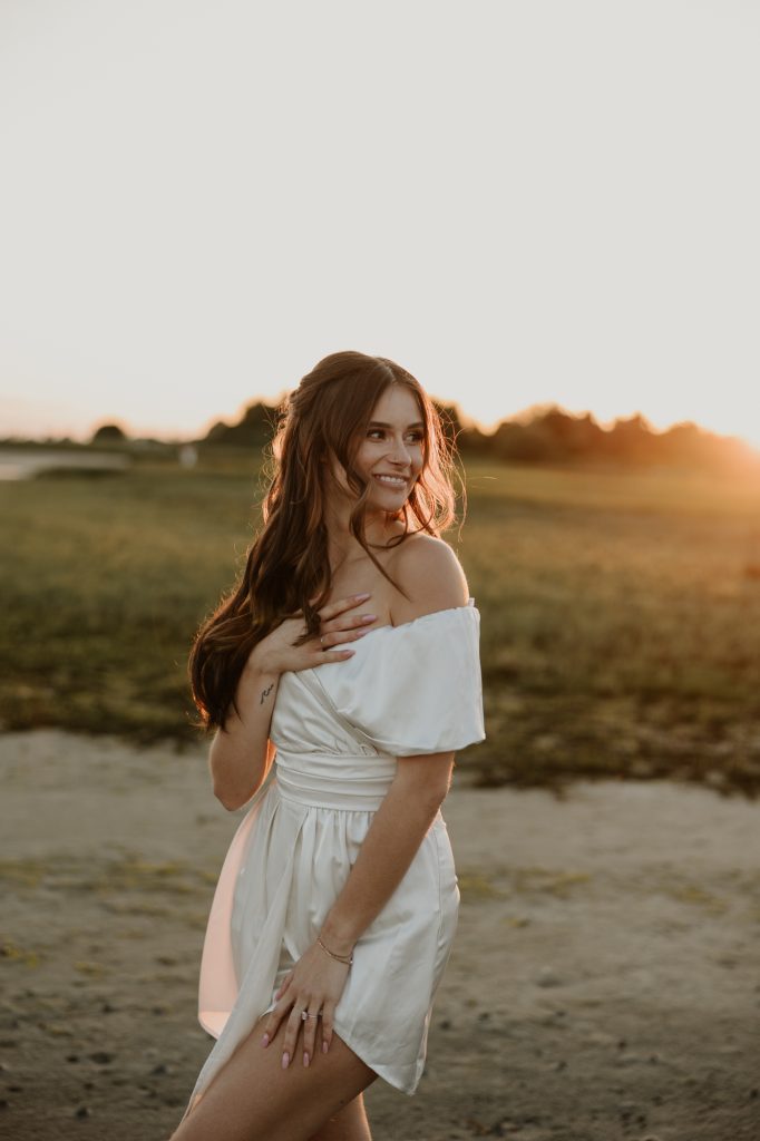 A woman with auburn hair in a white dress smiles at sunset on a grassy field