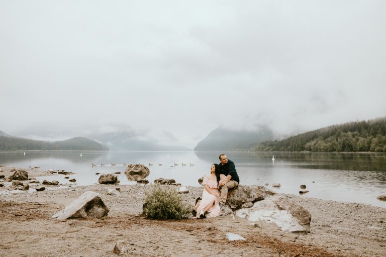 A couple sits together on a boulder at the edge of a misty mountain lake