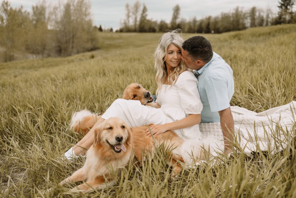 A couple cuddle in a grassy field with their golden retriever