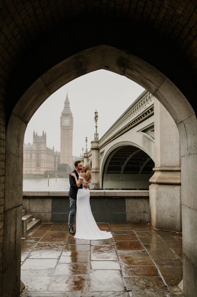 A couple stands beneath a stone archway with Big Ben across the river