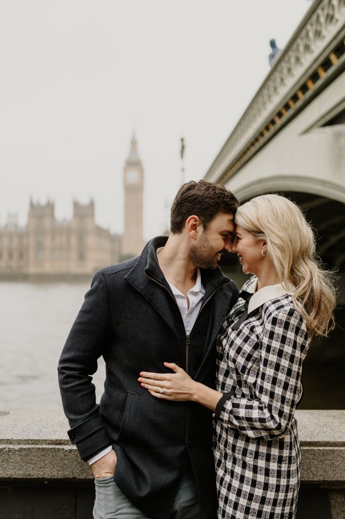 A couple lean in close beside a bridge on the Thames, with Big Ben in the distance