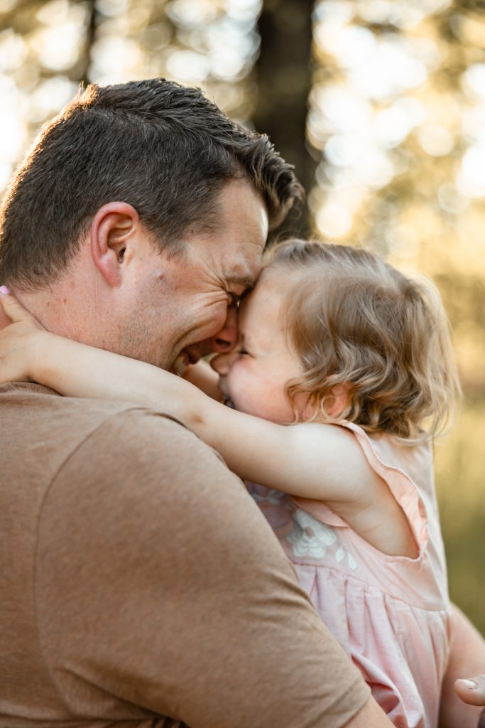 A dad and his toddler daughter touch noses, both laughing