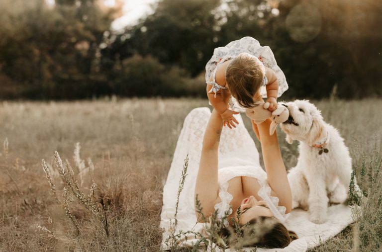 A mother lying in the long grass lifts her baby overhead while the family's fluffy white dog looks on
