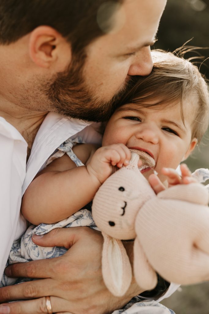 A dad kisses his baby's cheek as she clutches a knitted bunny
