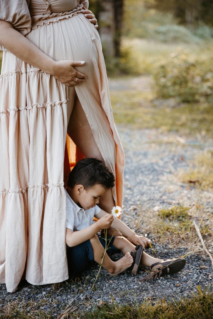 A little boy sits at his mother's feet holding a tiny flower, beside the hem of her long dress