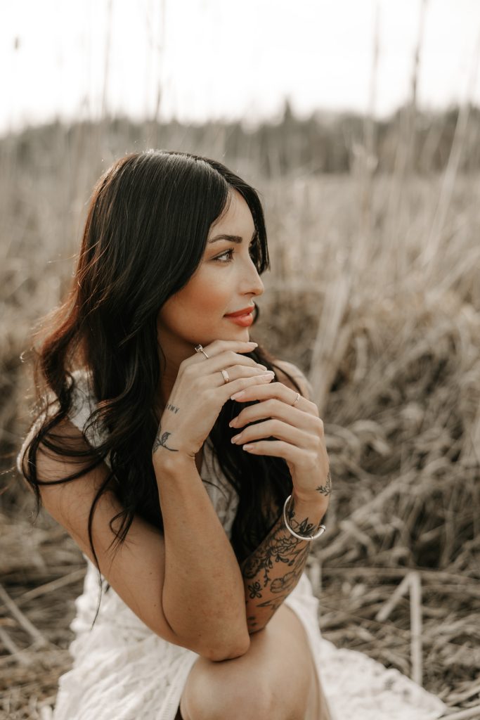 A woman with long dark hair sits in tall dry grass, her chin resting on her hand