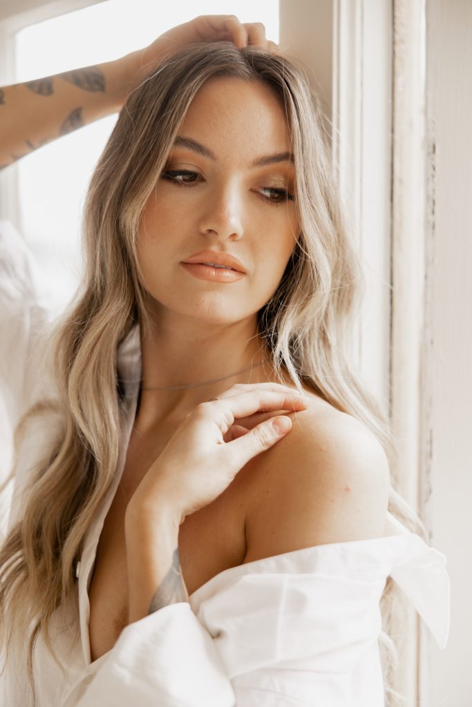 A woman in an off-the-shoulder white shirt looks down, a hand at her collarbone