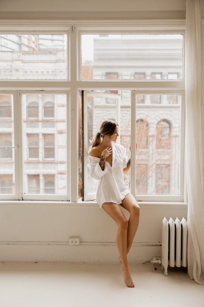 A woman in an oversized white shirt sits on the sill of a tall, sunlit loft window