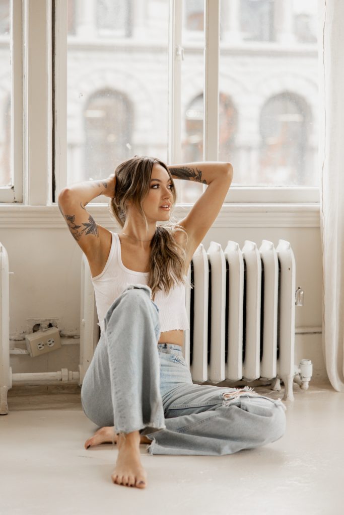 A woman in jeans sits on a loft floor by a radiator, hands in her hair