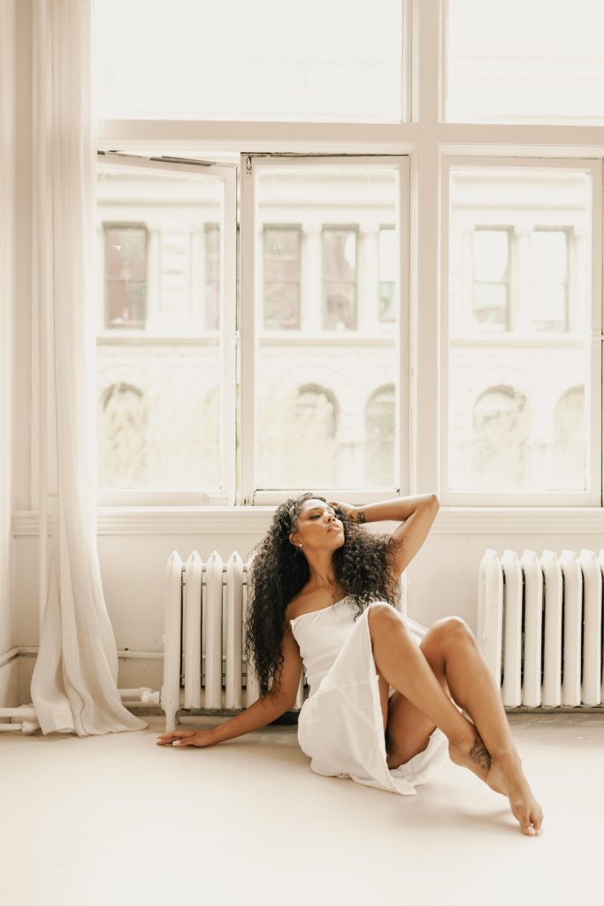 A woman in a white dress sits on the floor of a sunlit loft beneath tall windows