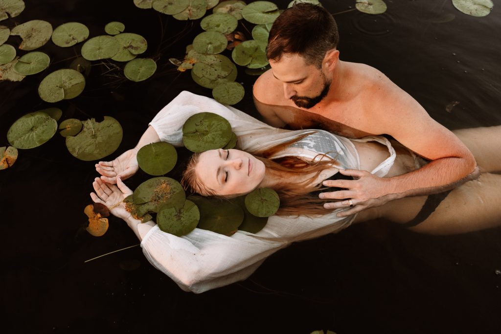 A couple float among lily pads in a lake, she leans back in his arms